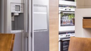 A stainless refrigerator with a water dispenser beside a column of built-in ovens in a modern kitchen