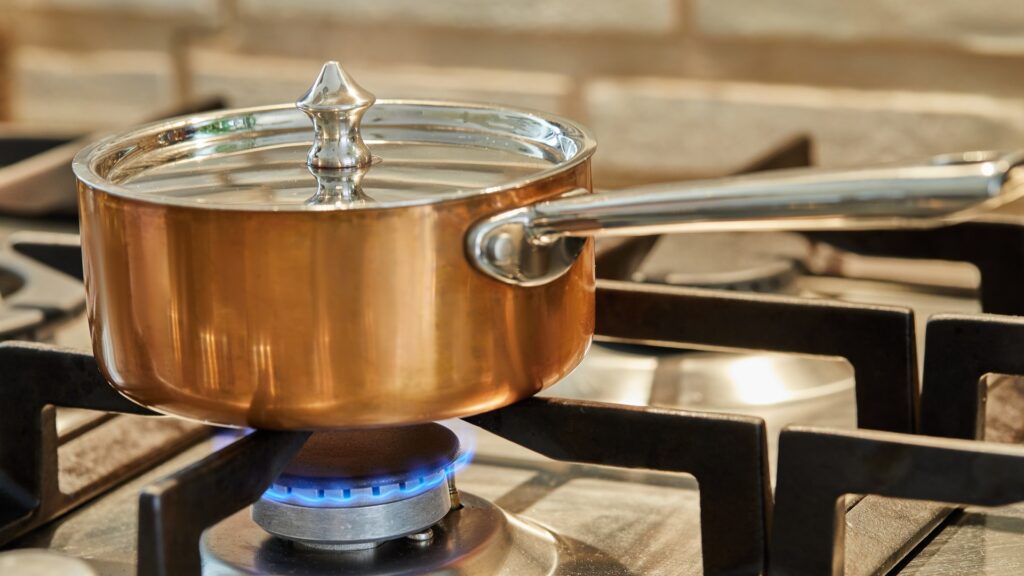 A covered saucepan sitting over a very low blue gas flame on a professional cooktop