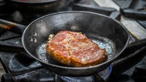 A steak searing in a cast iron skillet over a blue gas flame on a cooktop