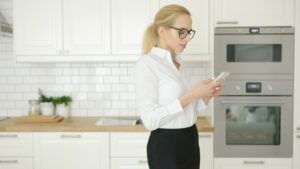A person standing with a phone beside a column of built-in ovens in a finished kitchen